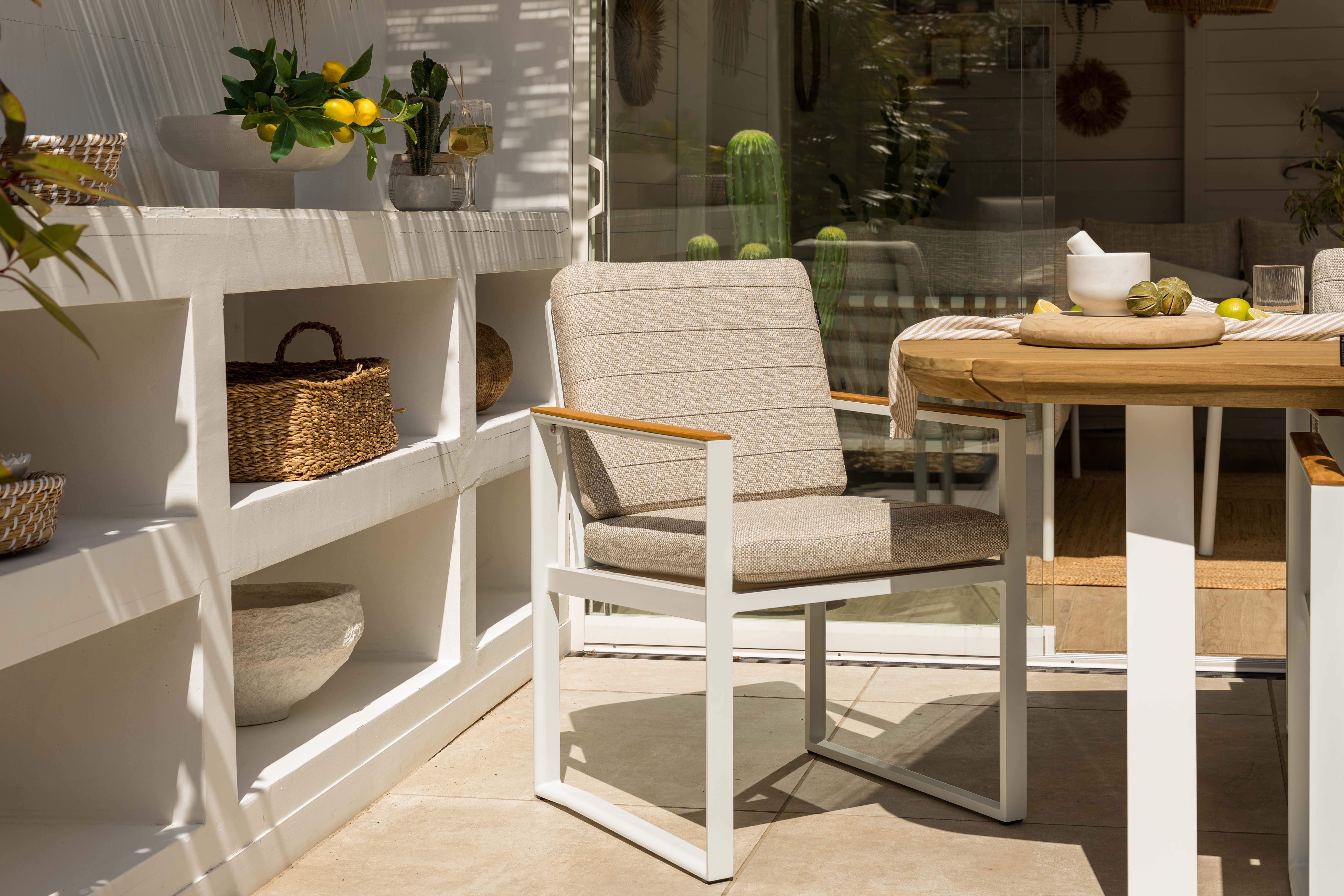 Geweven tuinstoel bij een houten tuintafel op een zonnig terras, met witte open kast en opbergmanden op de achtergrond.