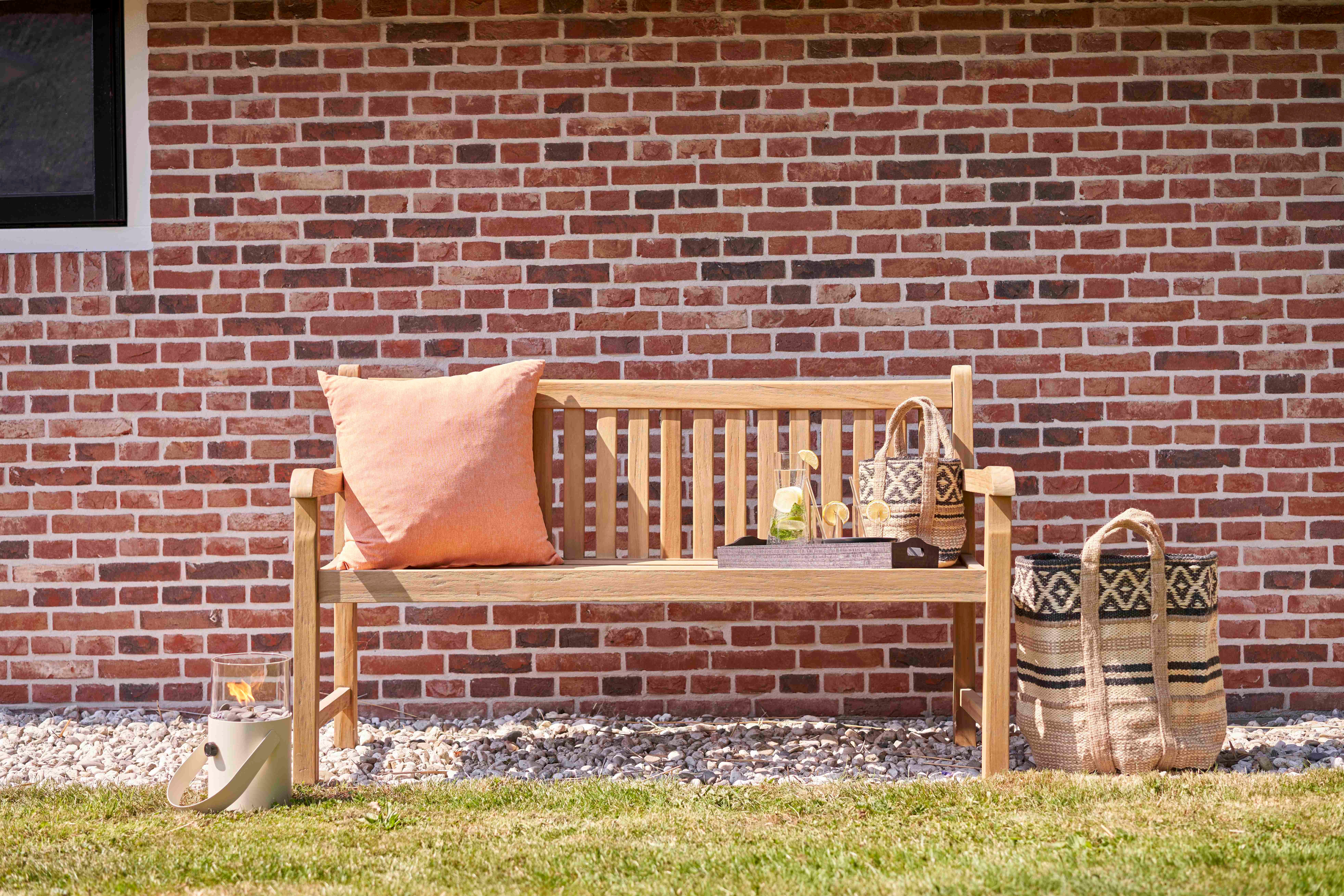 Klassieke teak parkbank van 150 cm met oranje kussen tegen een rode bakstenen muur.