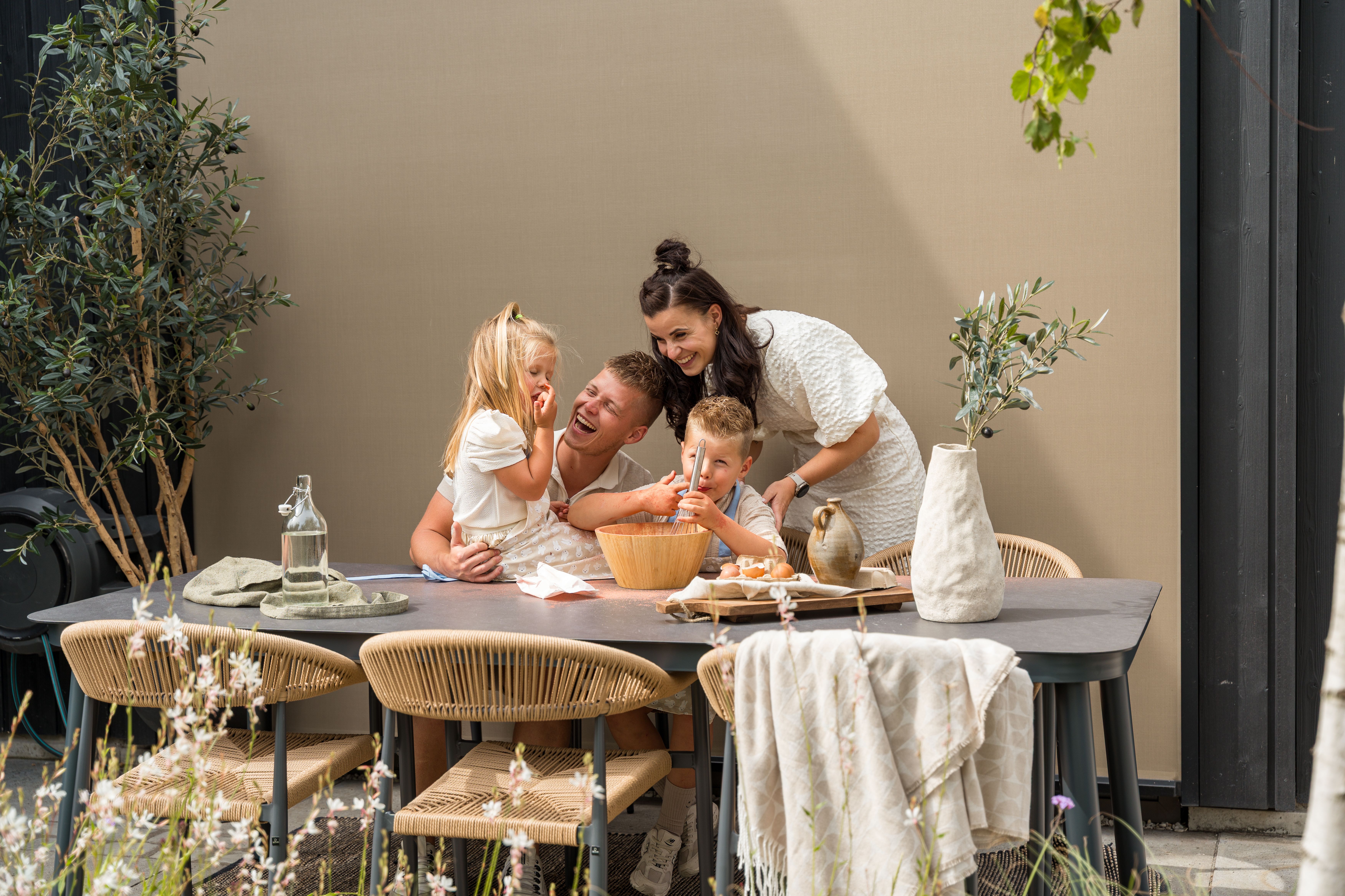 Gezin met twee jonge kinderen zit lachend aan een buitentafel met rieten stoelen in een zonnige tuin, terwijl ze samen fruit eten en genieten van een ontspannen moment op het terras.