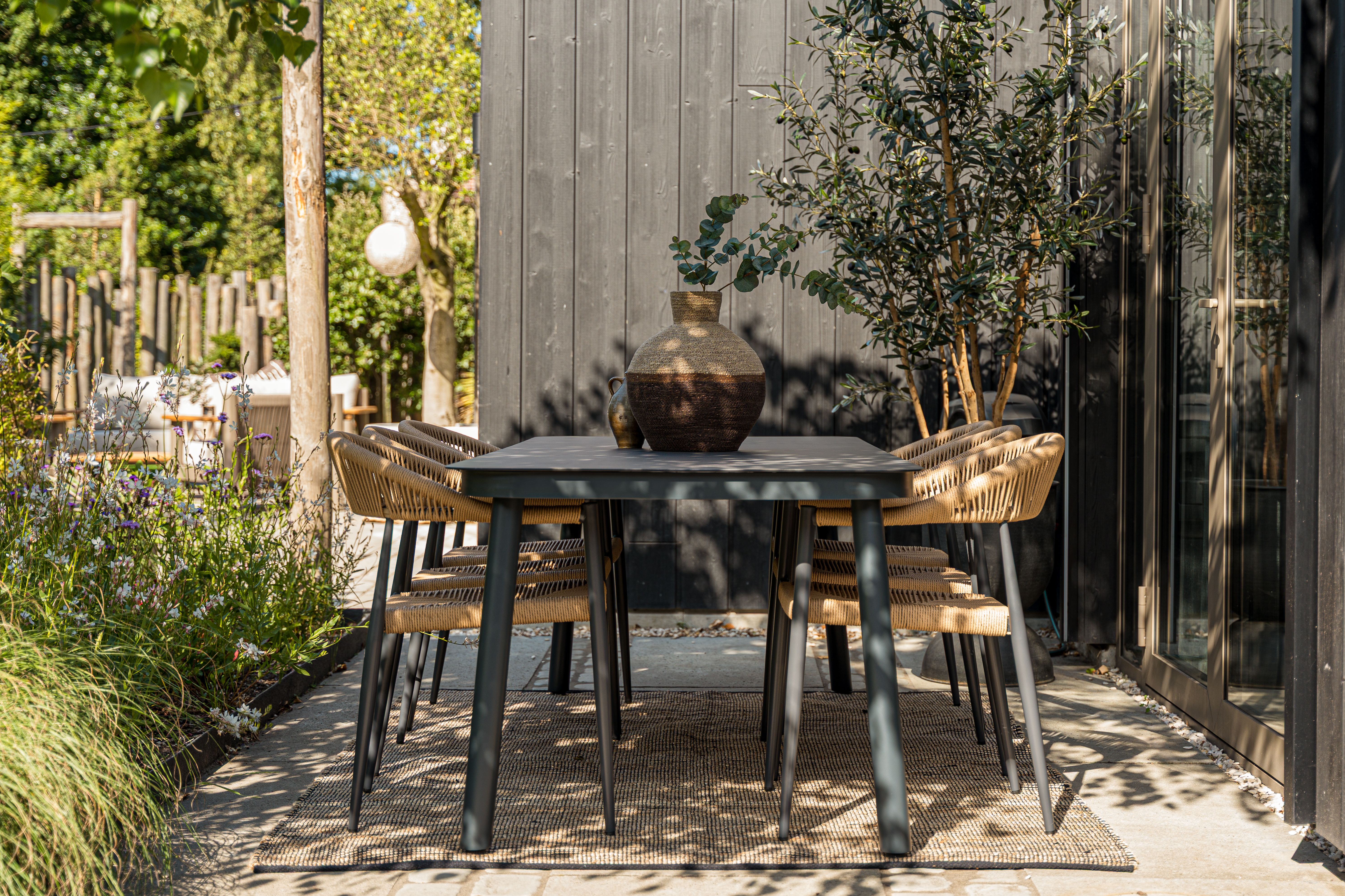 Moderne buitentafel met zwart blad en rieten stoelen op een houten terras naast een donkere gevel, omringd door bloeiende tuinplanten.