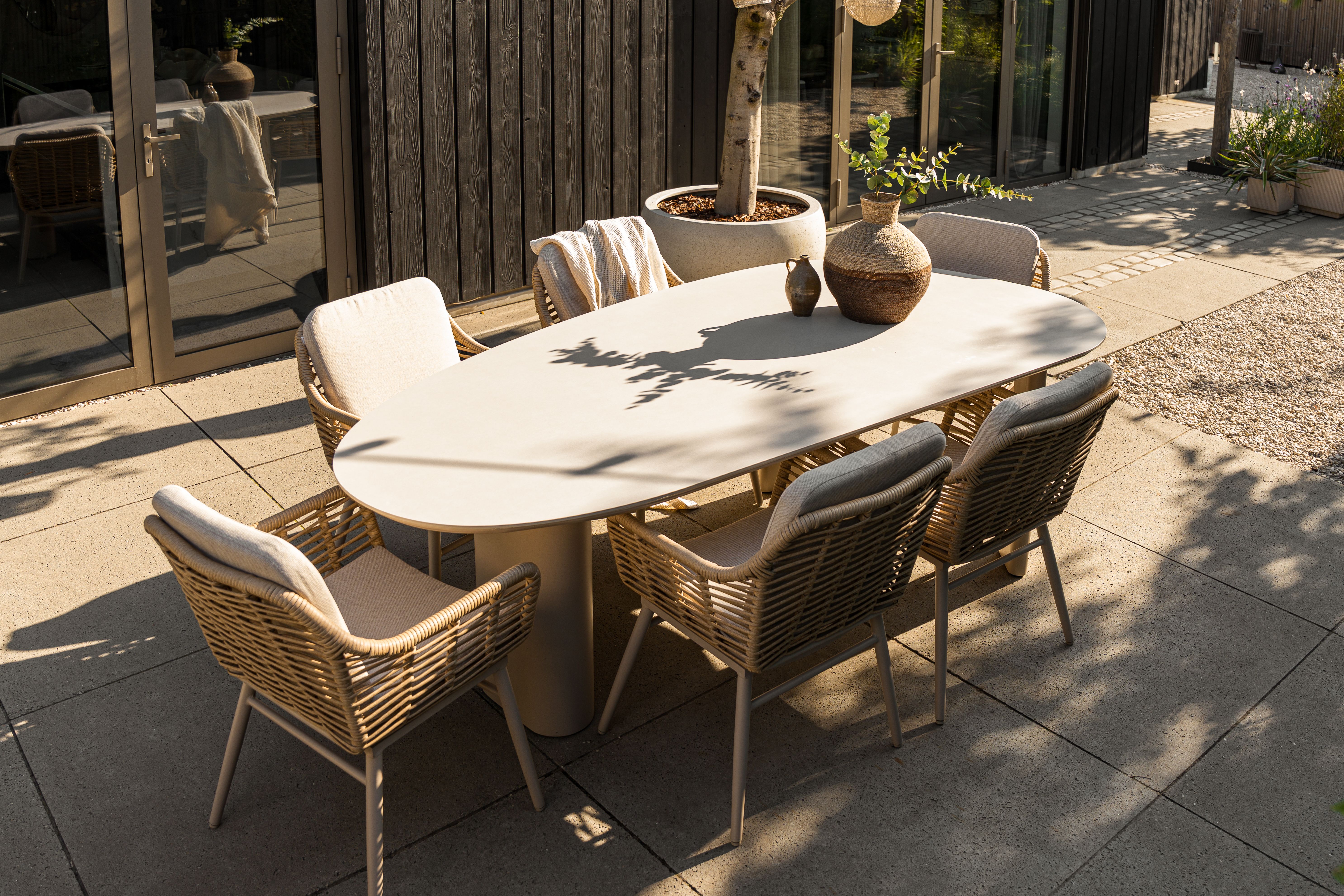 Moderne tuinset met organische eettafel in zand kleur en zes wicker tuinstoelen met beige kussens op een zonnig terras bij een houten gevel.