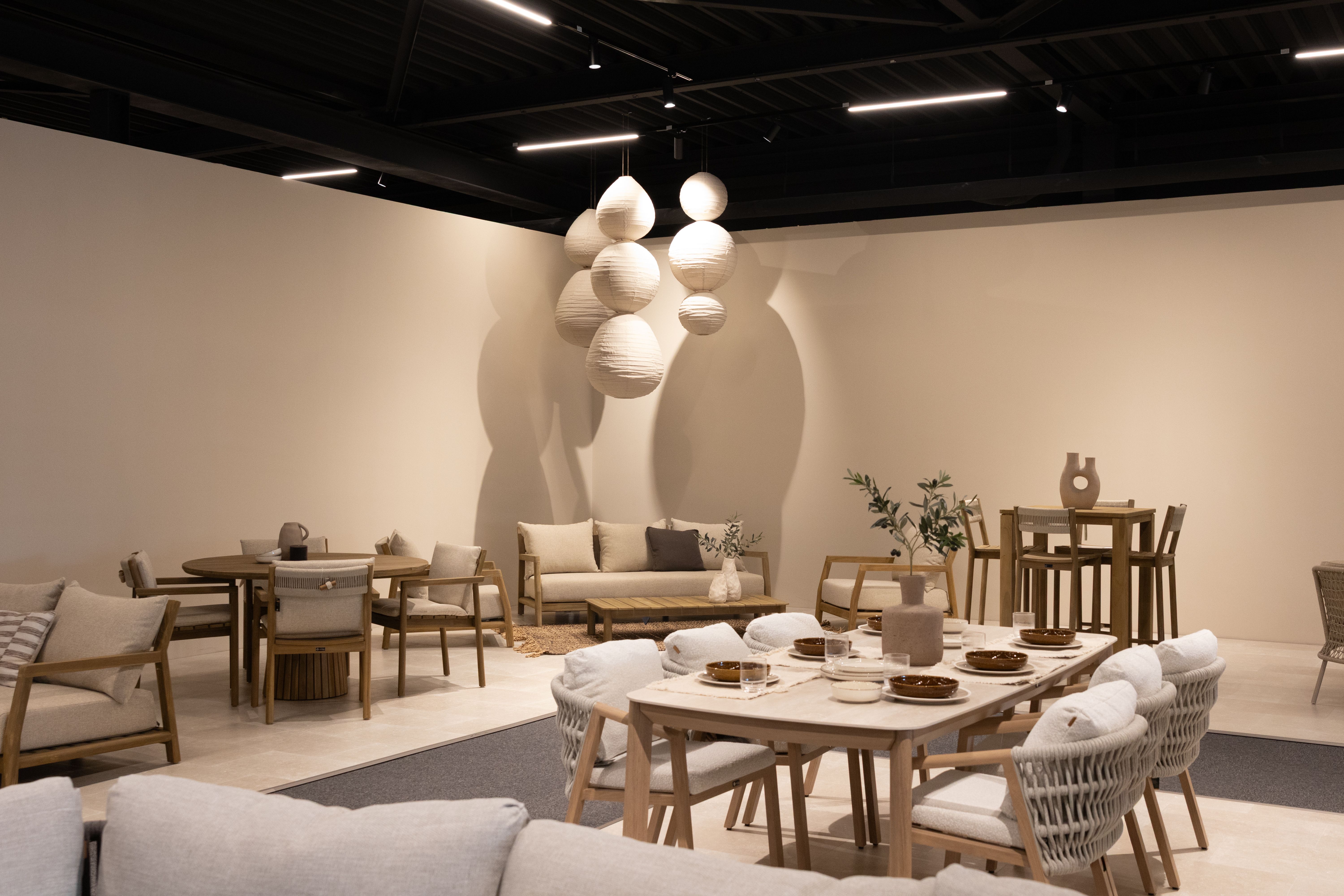 Showroom met moderne houten tuinset: eettafel met stoelen en loungeset in neutrale tinten, sfeervol verlicht met ronde hanglampen.