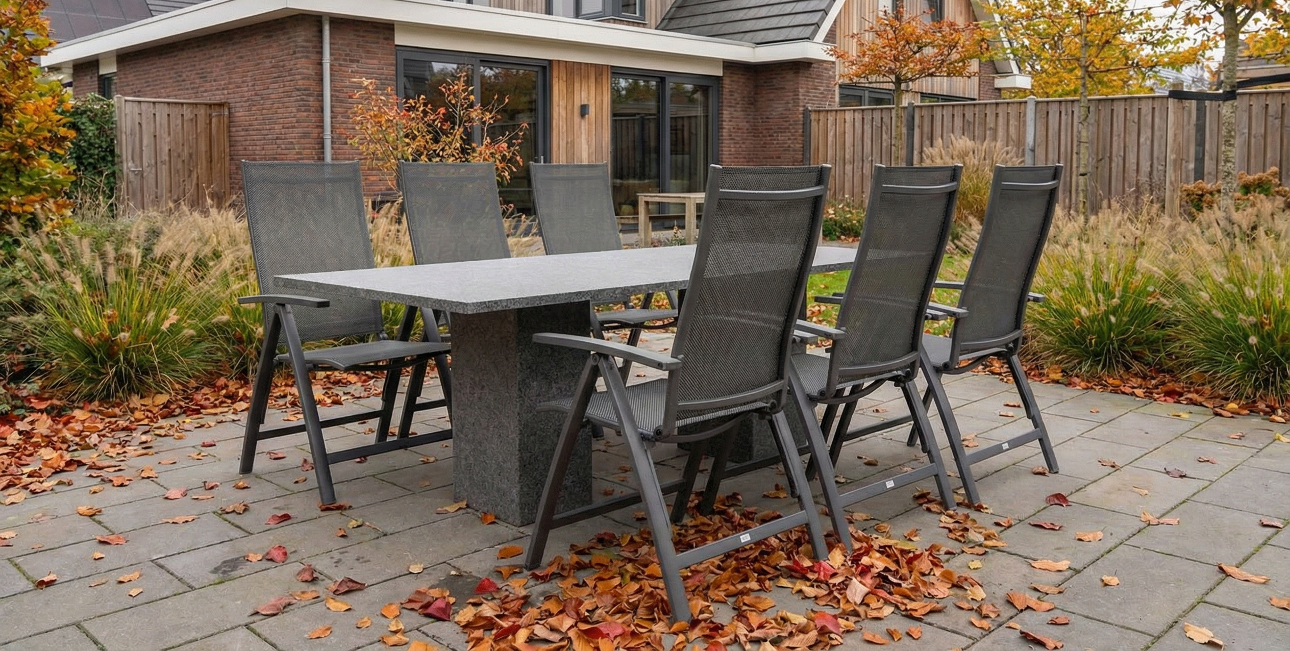 Tuinset met granieten tuintafel en verstelbare tuinstoelen van textileen op een betegeld terras, omringd door beplanting met herfstbladeren en een herfstachtige sfeer.