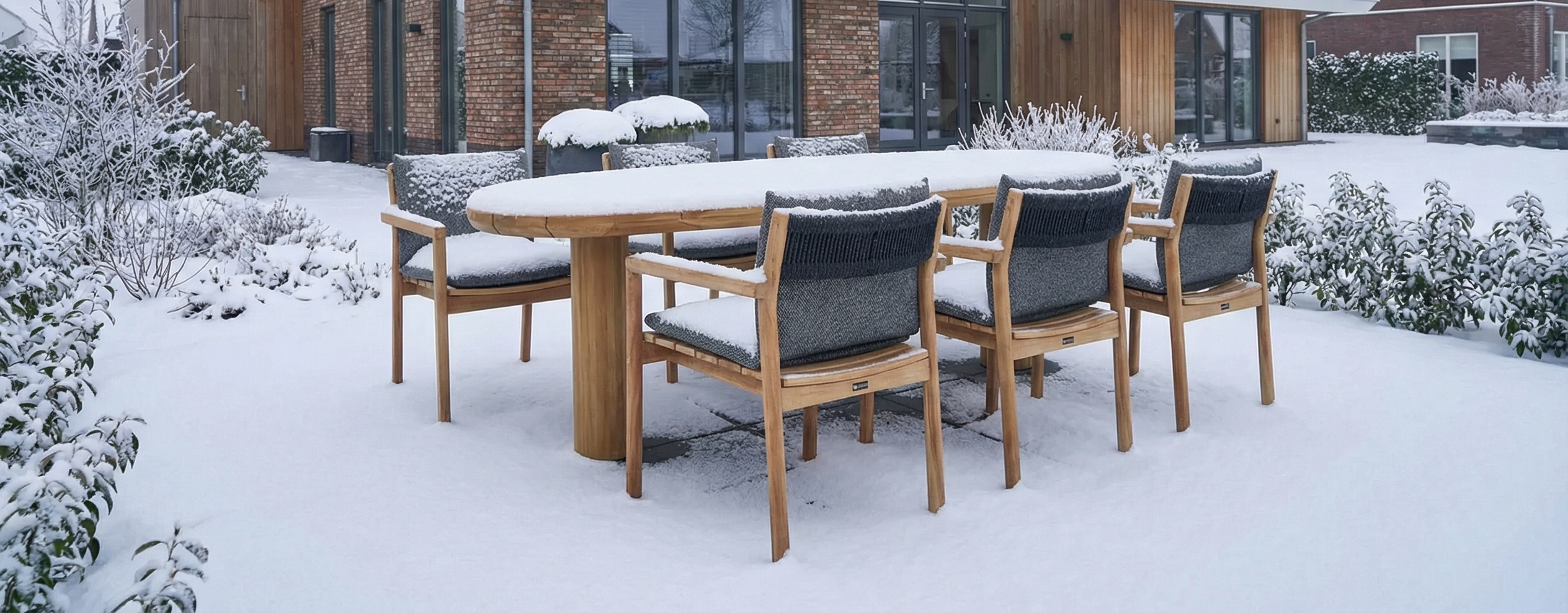 Teakhouten tuinset met robuuste teakhouten tuintafel en bijpassende teakhouten tuinstoelen met donkere kussens, geplaatst in een besneeuwde tuin bij een moderne woning.
