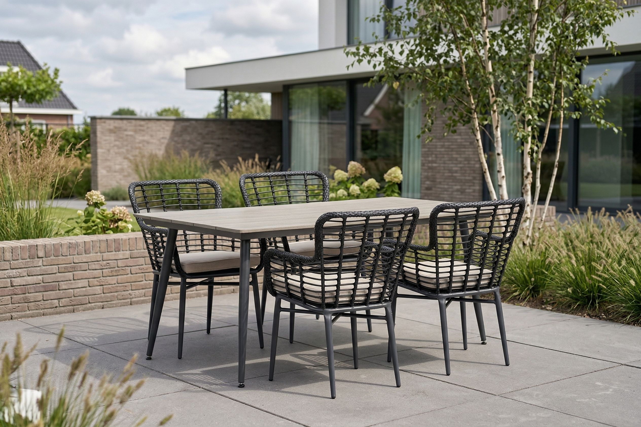 Moderne tuinset met rechthoekige tuintafel en vier zwarte wicker tuinstoelen met lichte kussens op een strak terras bij een moderne woning met groene tuin.