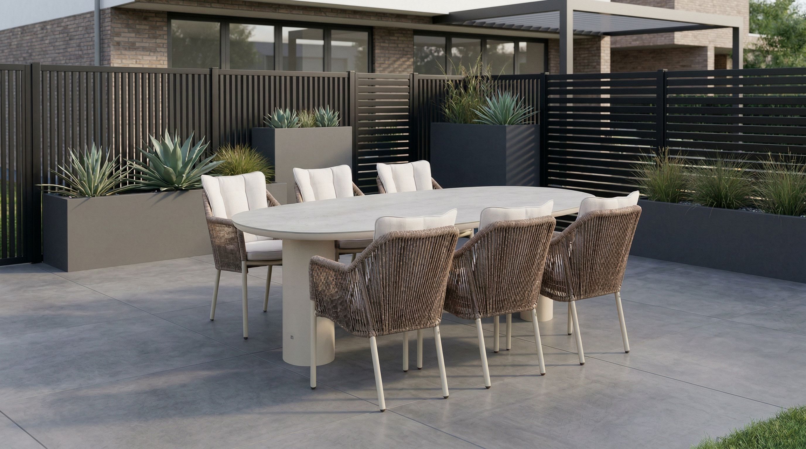 Wicker tuinset met ovale tuintafel en zes rope tuinstoelen met lichte kussens op een ruim terras, geplaatst in een groene tuin met siergrassen en bomen.