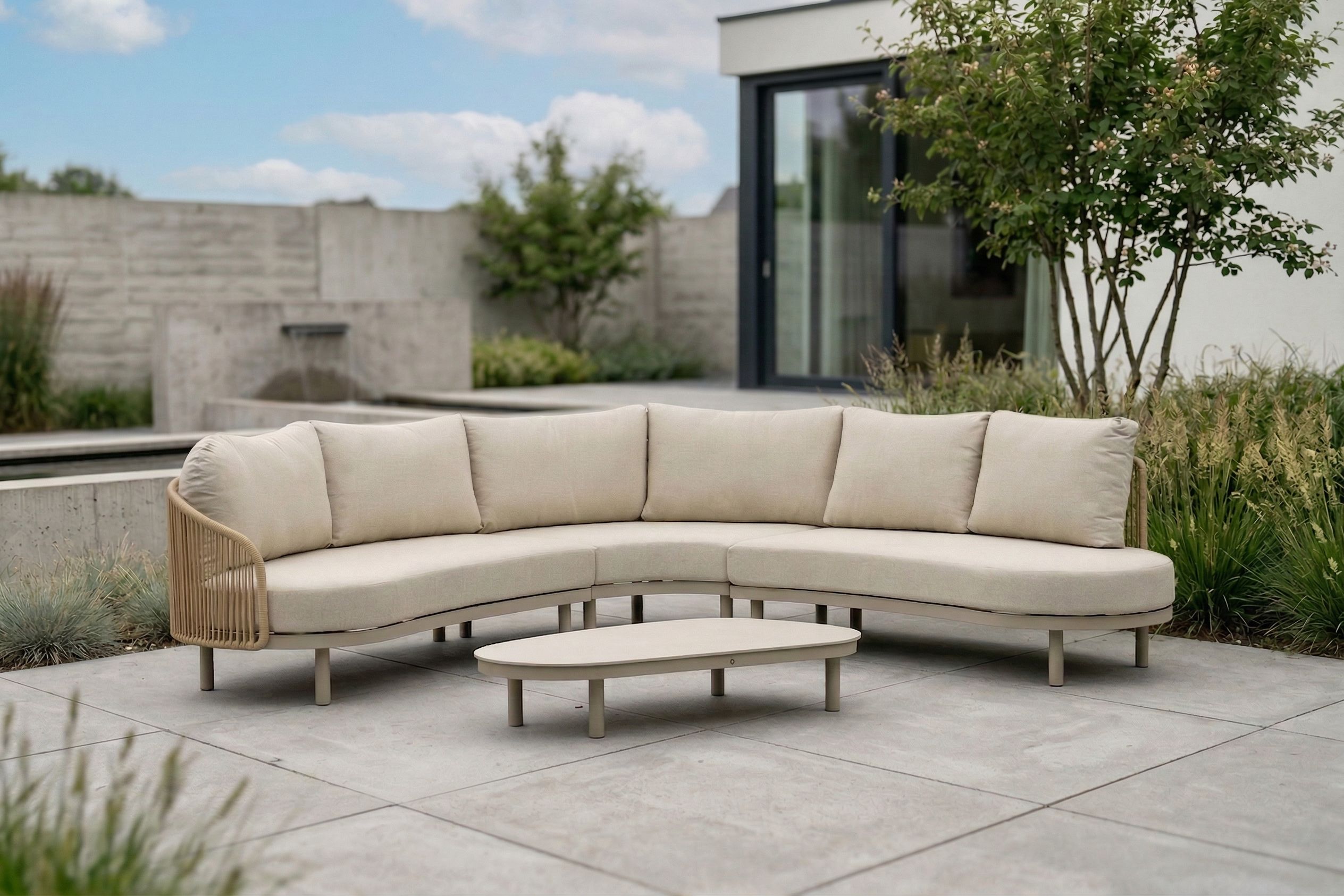 Luxe ronde loungeset met beige kussens en lage loungetafel op een ruim terras, geplaatst in een moderne tuin met siergrassen en een stijlvolle woning op de achtergrond.