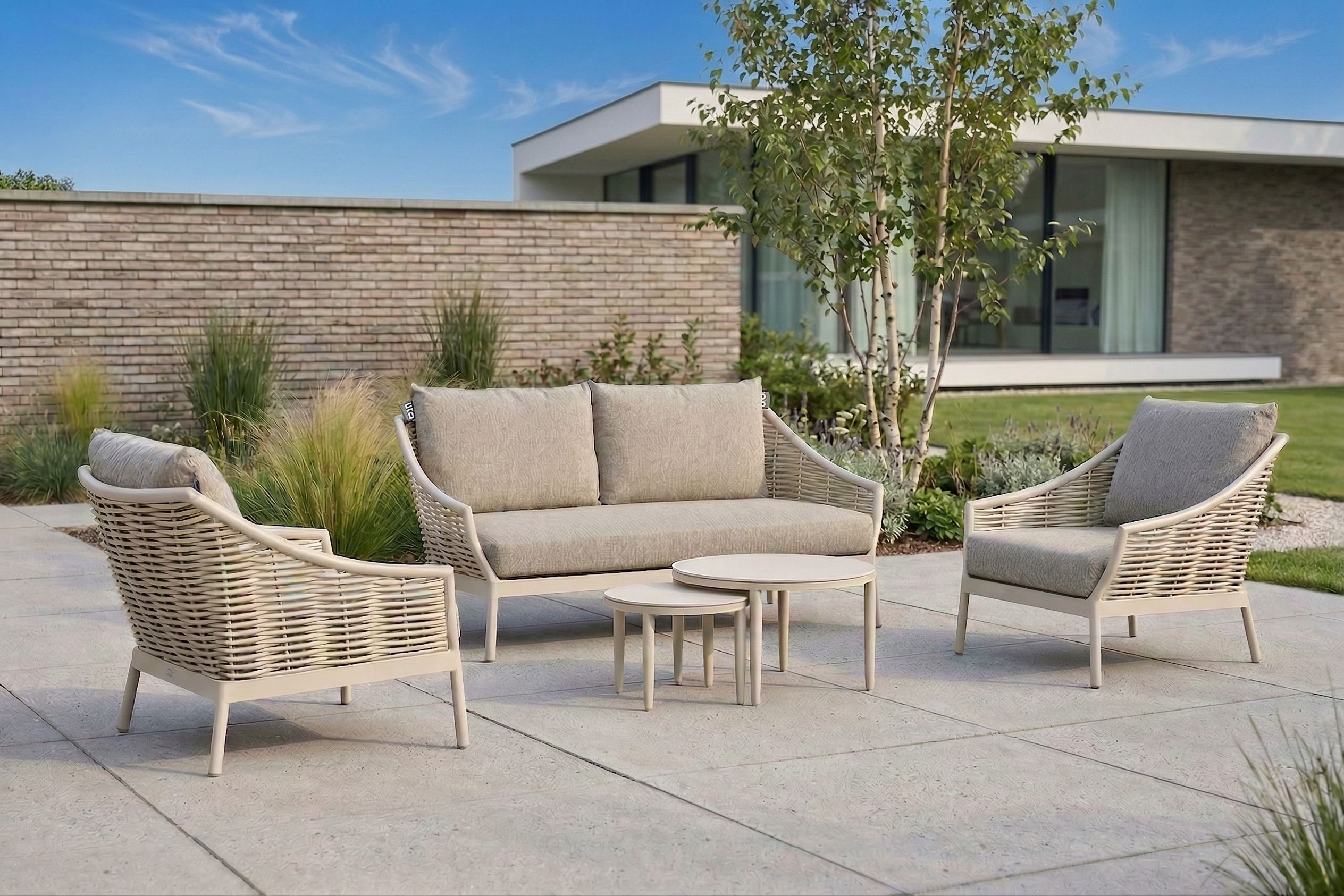 Een moderne beige wicker loungeset op een betegeld terras, bestaande uit een driezitsbank, twee loungestoelen met dikke kussens en twee ronde bijzettafels.