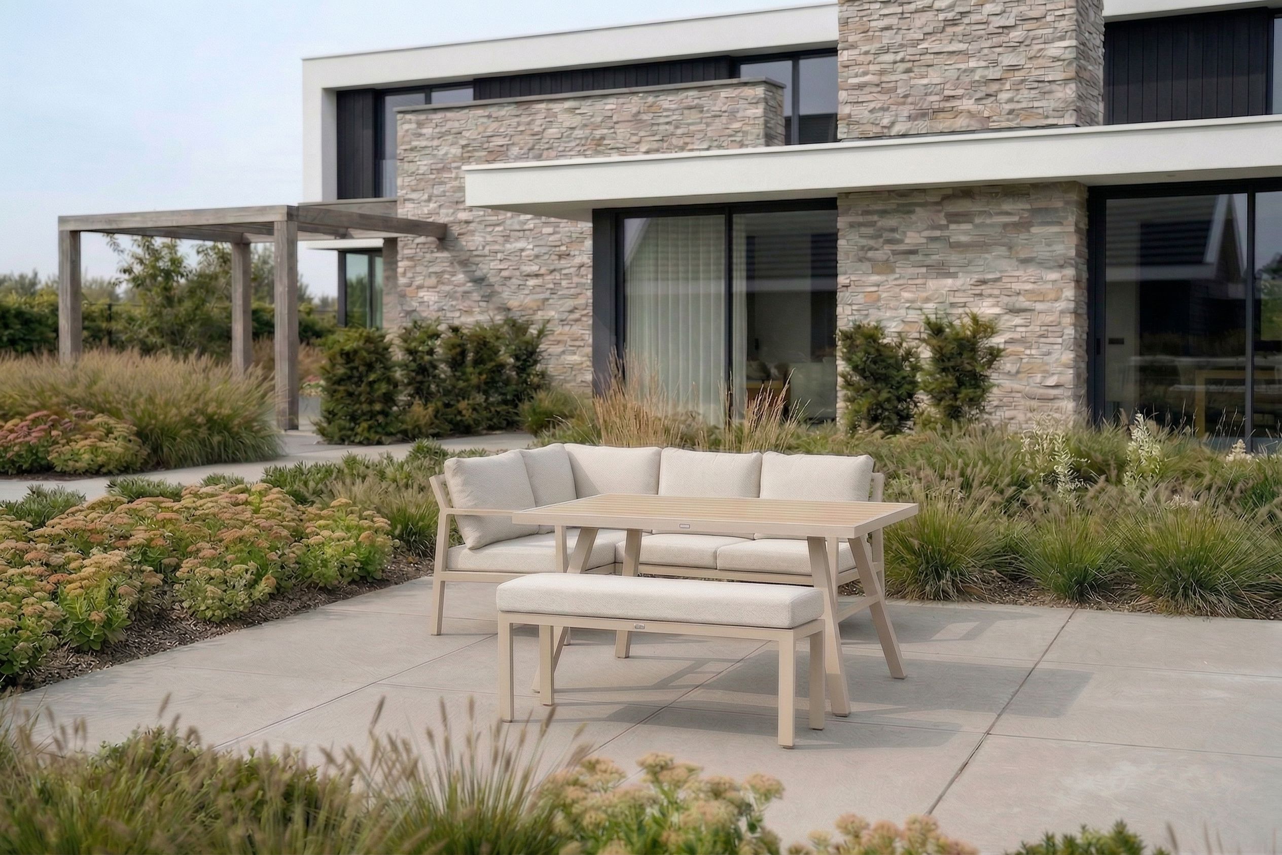 Beige hoek loungeset met kussens, lounge- diningtafel en bijpassende picknickbank op een modern terras bij een woning met stenen gevel en groene tuin.