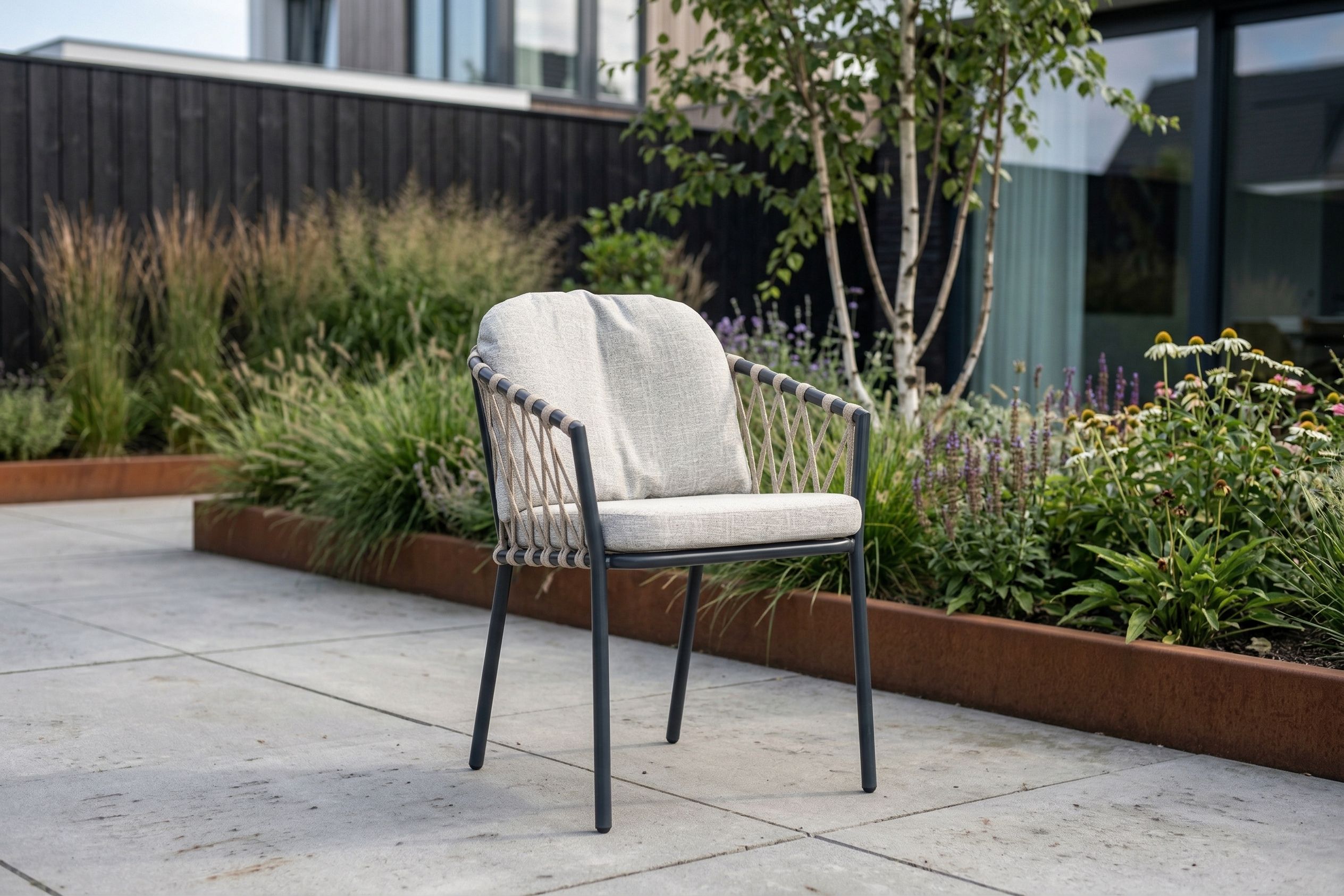 Moderne rope tuinstoel met beige kussen naast een kleine bijzettafel op een houten terras, omringd door groene planten bij een moderne woning.