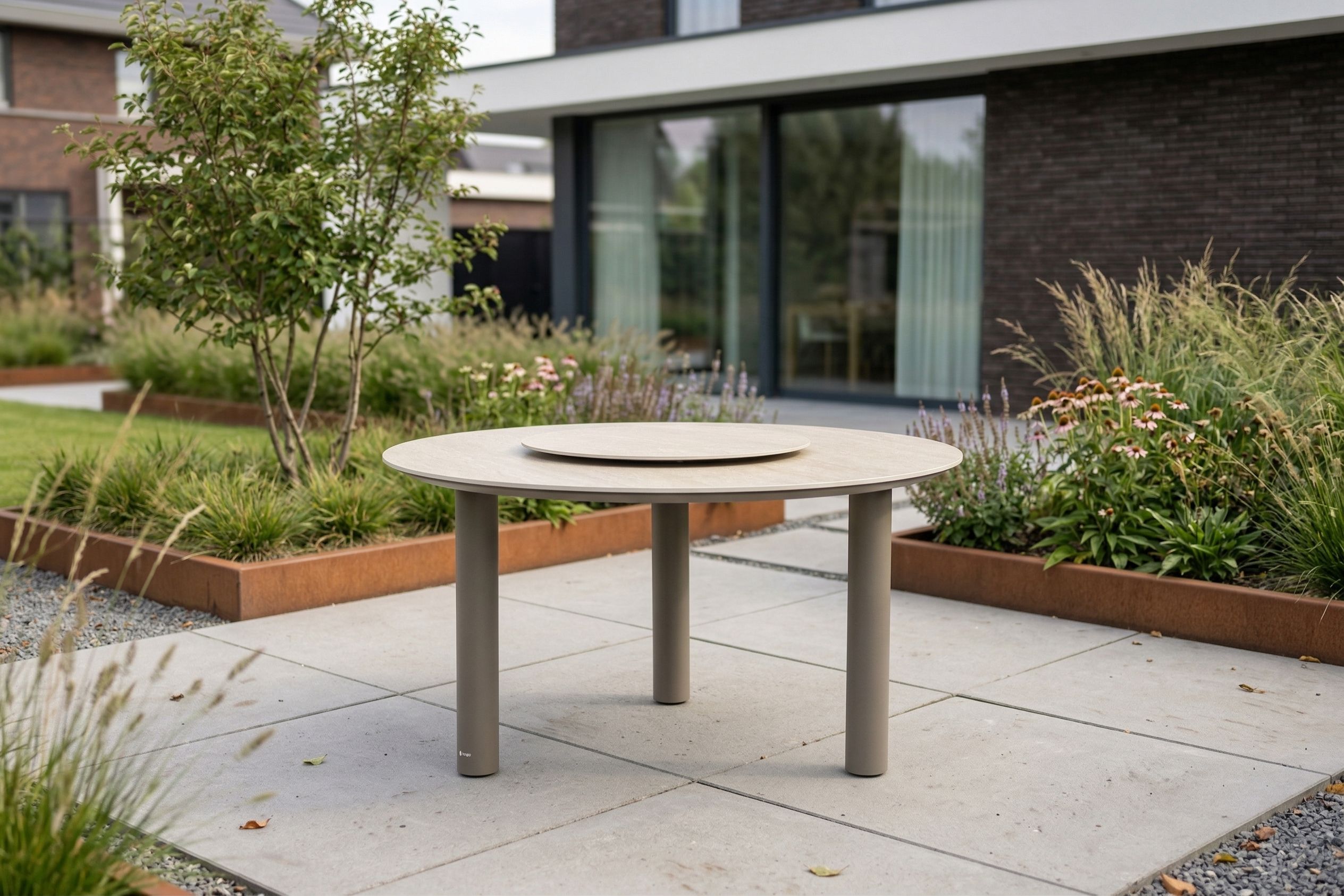 Moderne ovale tuintafel in loft kleur met drie ronde tafelpoten op een strak terras, geplaatst in een stijlvolle tuin met siergrassen en een zwarte schutting.