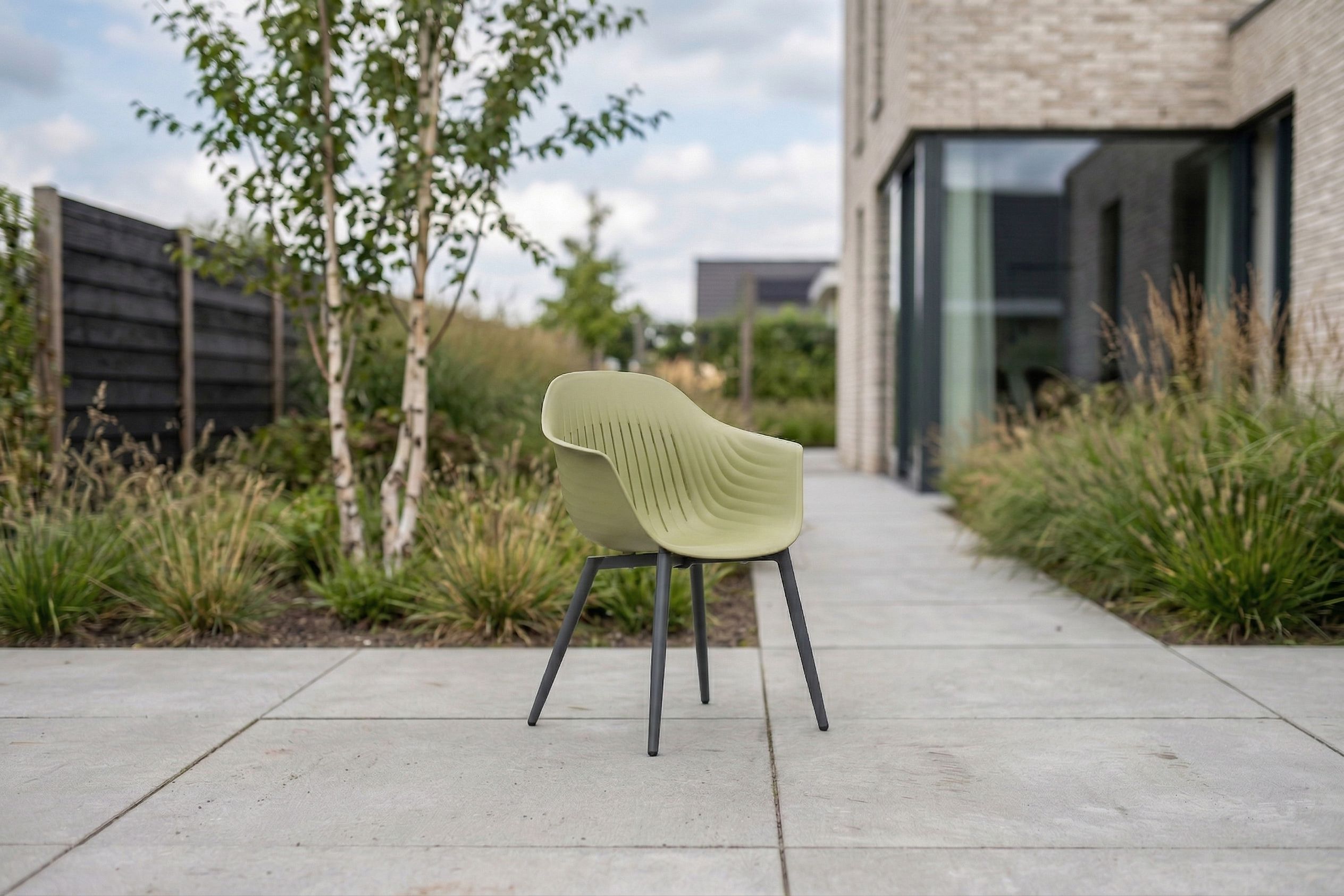 Groene kunststof tuinstoel met armleuningen en zwart metalen poten op een modern terras, omringd door siergrassen en een strakke woning.