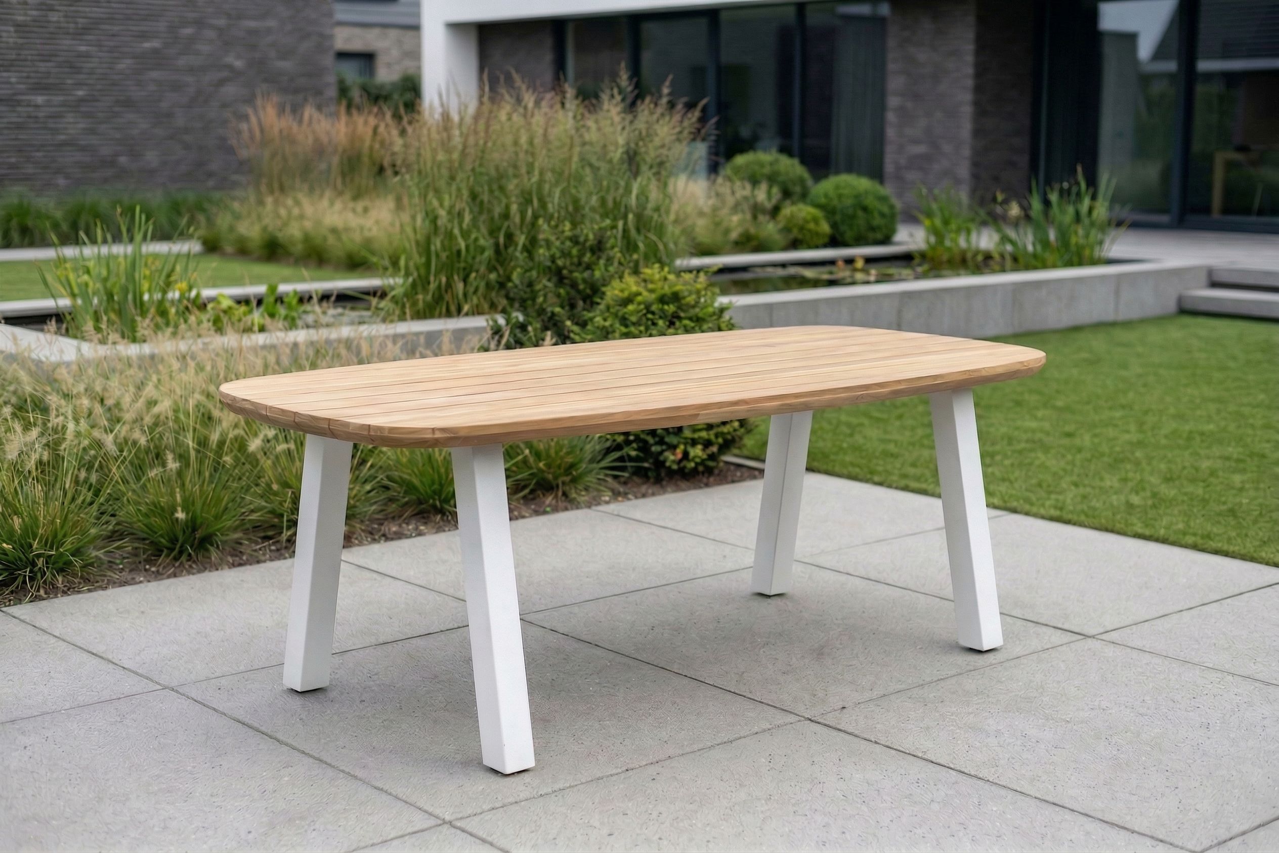 Een organische tuintafel met een lichtbruin houten blad en vier schuingeplaatste witte poten, staand op grote grijze terrastegels met een groene tuin op de achtergrond