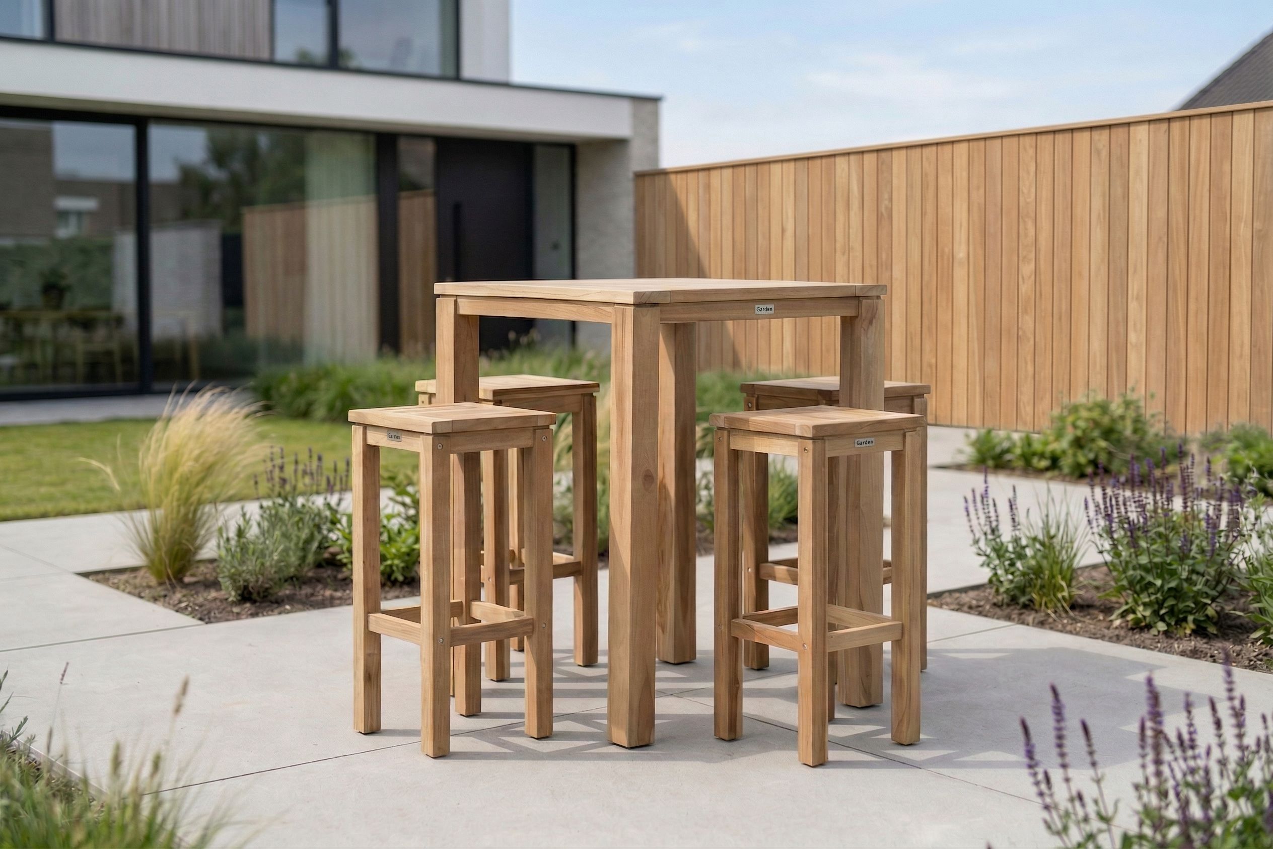Houten bartafel met bijpassende krukken op een modern terras, geplaatst in een strak aangelegde tuin met sierbeplanting en teakhouten schutting.