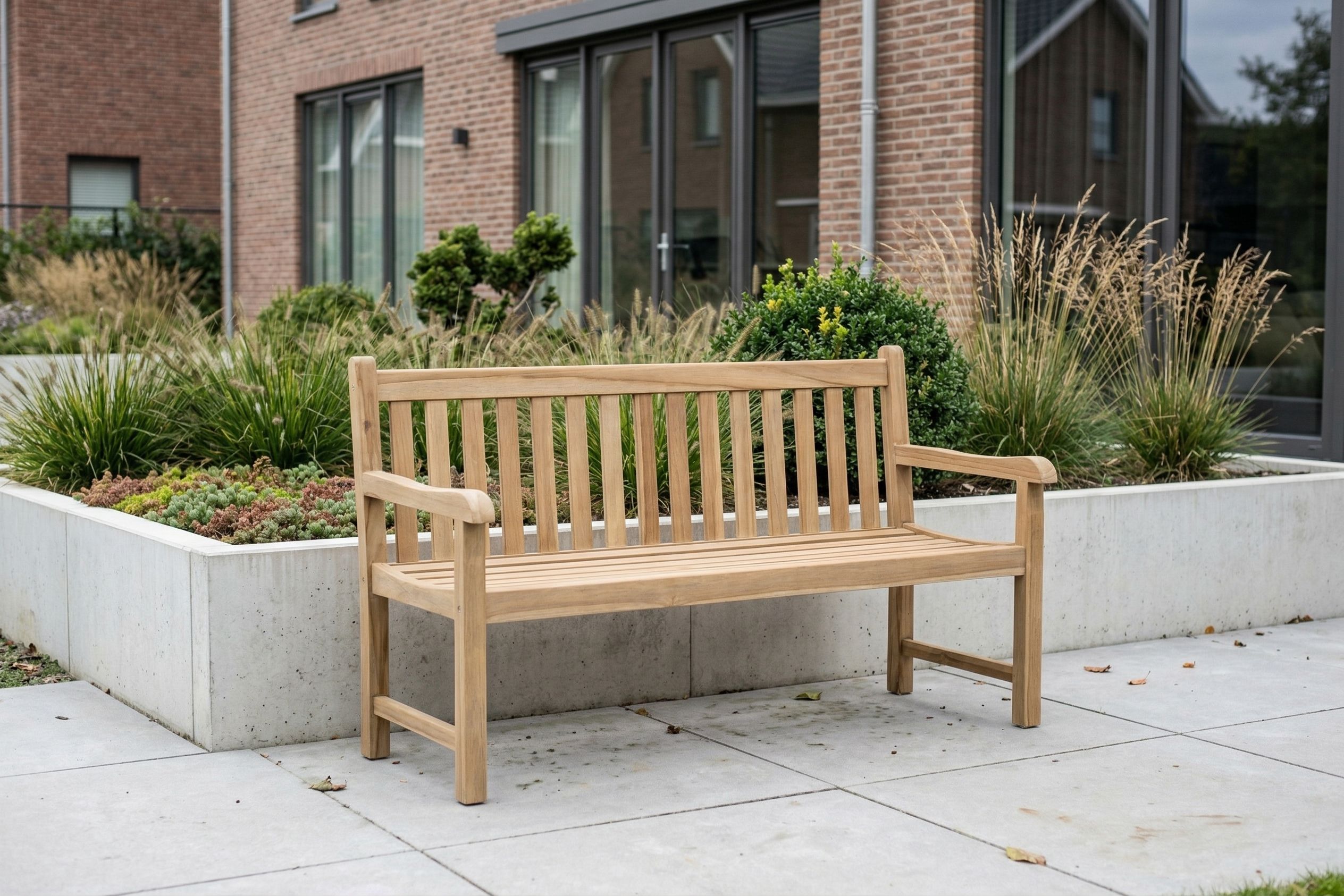 Teakhouten tuinbank met rugleuning en armleuningen op een modern terras, geplaatst voor een groene border bij een woning met bakstenen gevel.