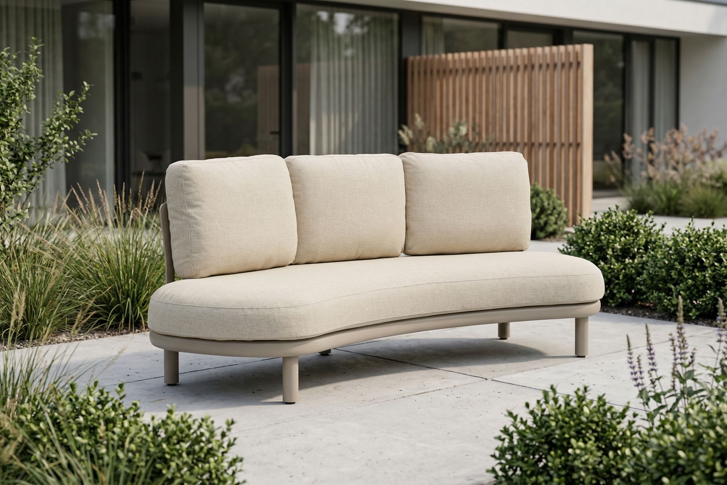 Beige design loungebank met afgeronde vormen en drie kussens op een modern terras, omringd door groene beplanting en een strakke woning.