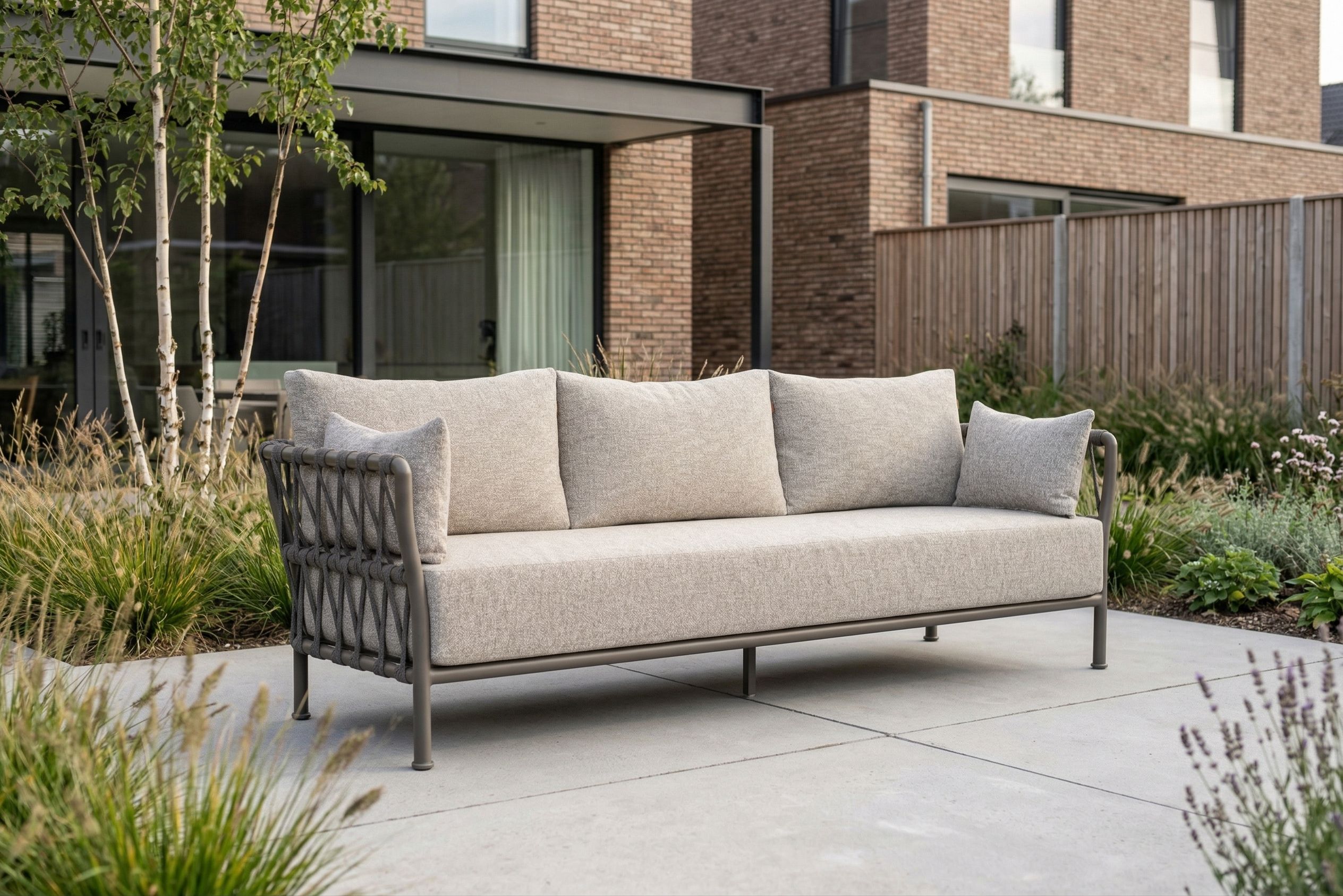 Moderne 3-zits loungebank met lichtgrijze kussens en rope zijkanten op een strak terras, geplaatst in een groene tuin bij een bakstenen woning.