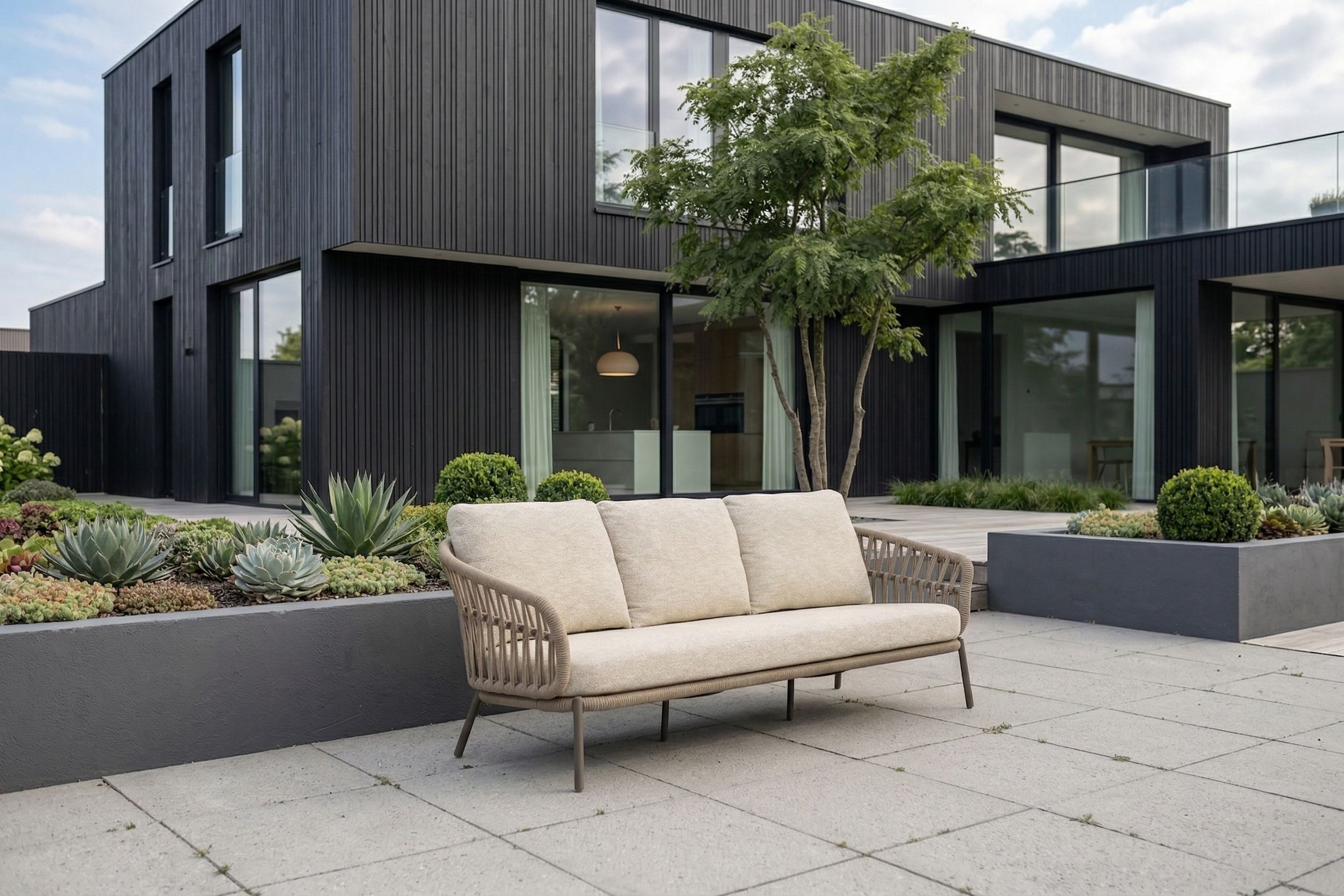 Moderne luxe tuinbank met beige kussens op strak aangelegd terras bij een minimalistische zwarte woning met grote ramen en groene beplanting.