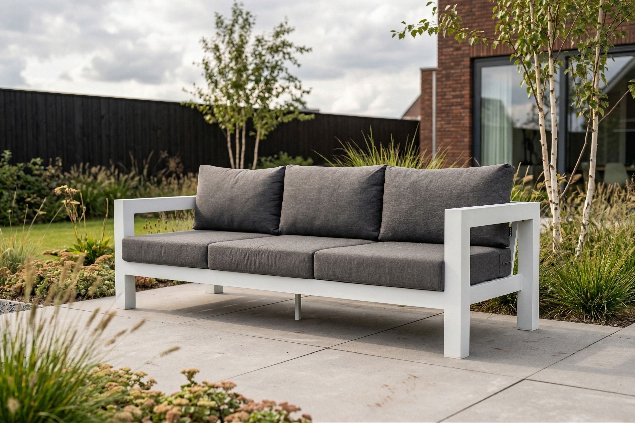 Moderne 3-zits aluminium loungebank in het wit met donkergrijze kussens op een betegeld terras in de achtertuin.