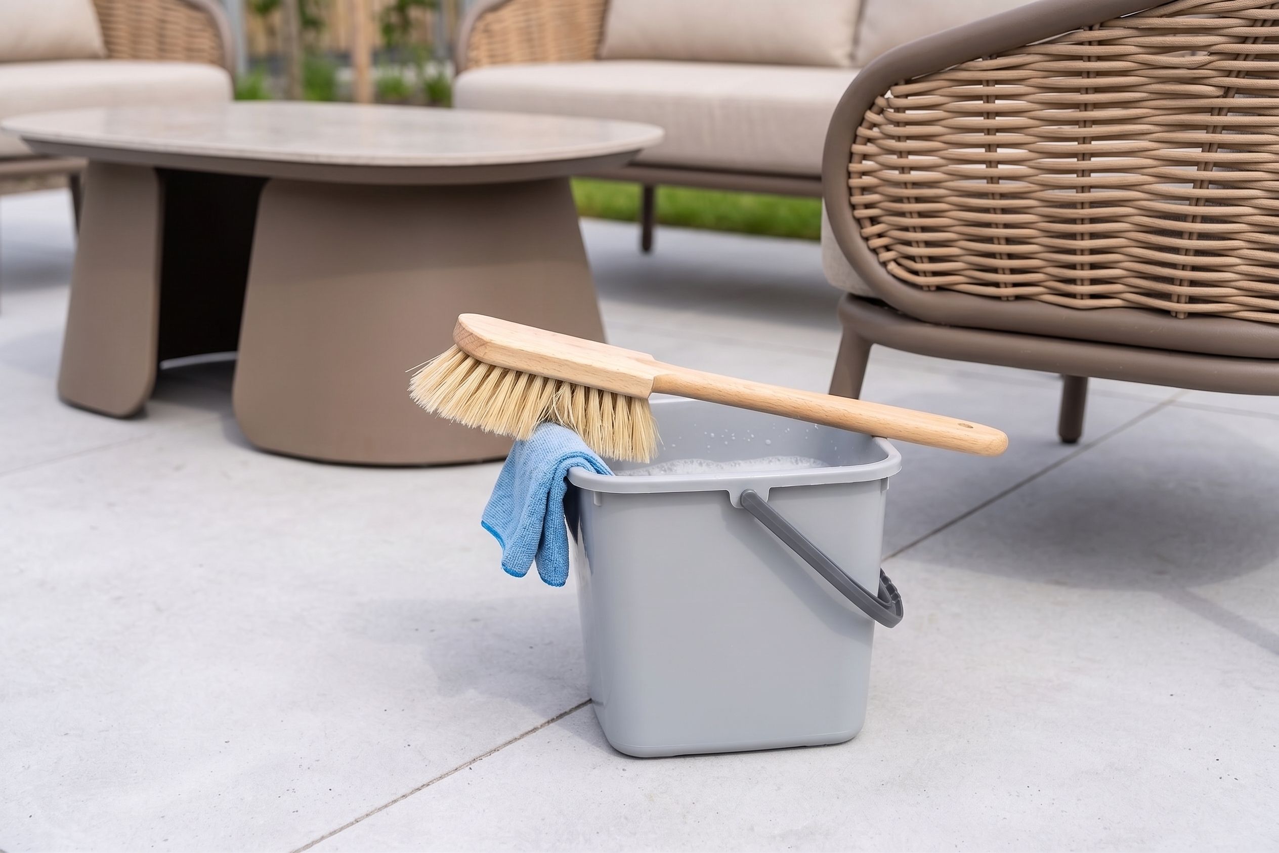 Een grijze emmer met sop, een blauw schoonmaakdoekje en een houten wasborstel staan op een terras naast een wicker loungeset.