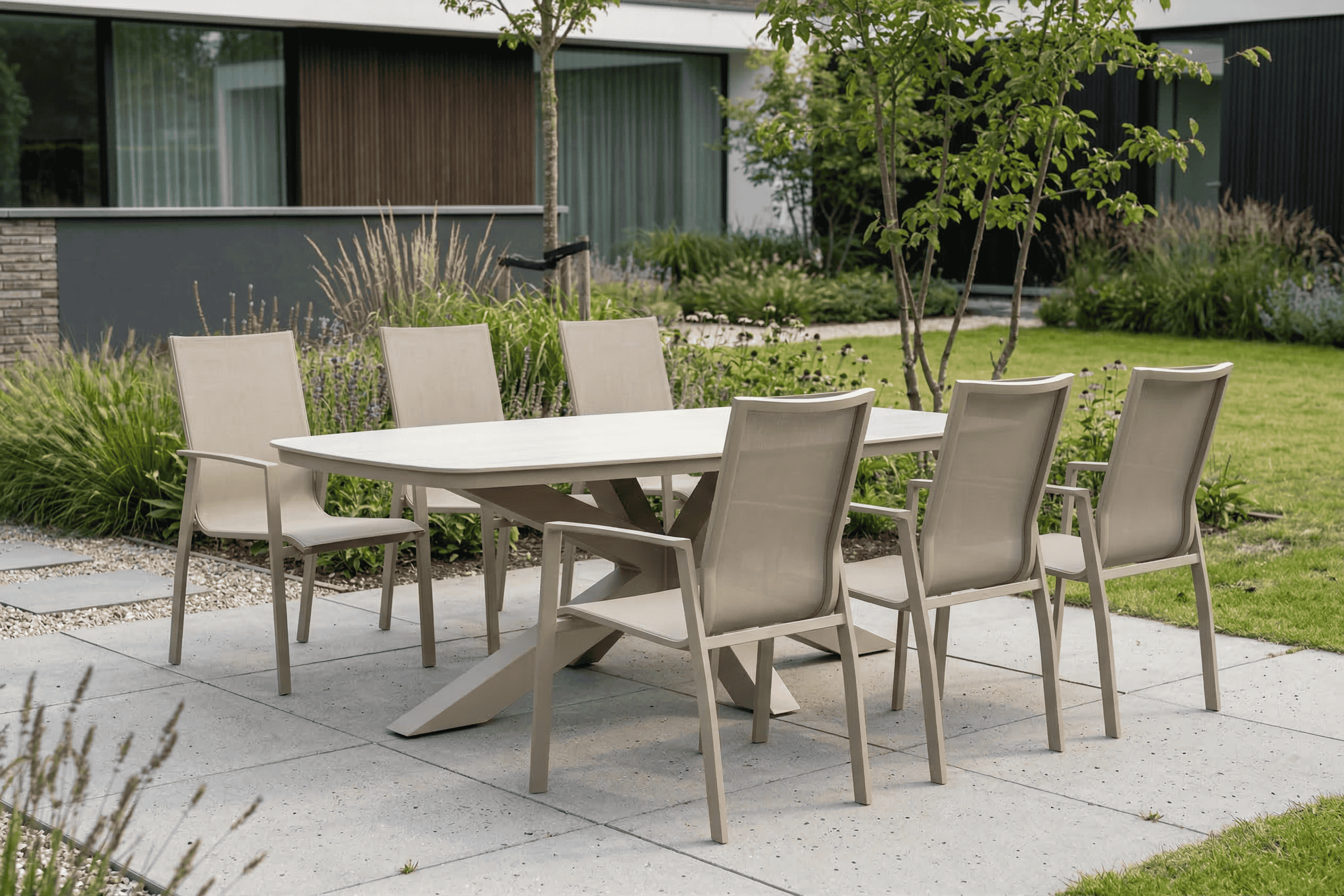 Aluminium tuinset met ovale tuintafel en zes beige tuinstoelen op een modern terras, geplaatst in een groene tuin met siergrassen en beplanting bij een moderne woning.