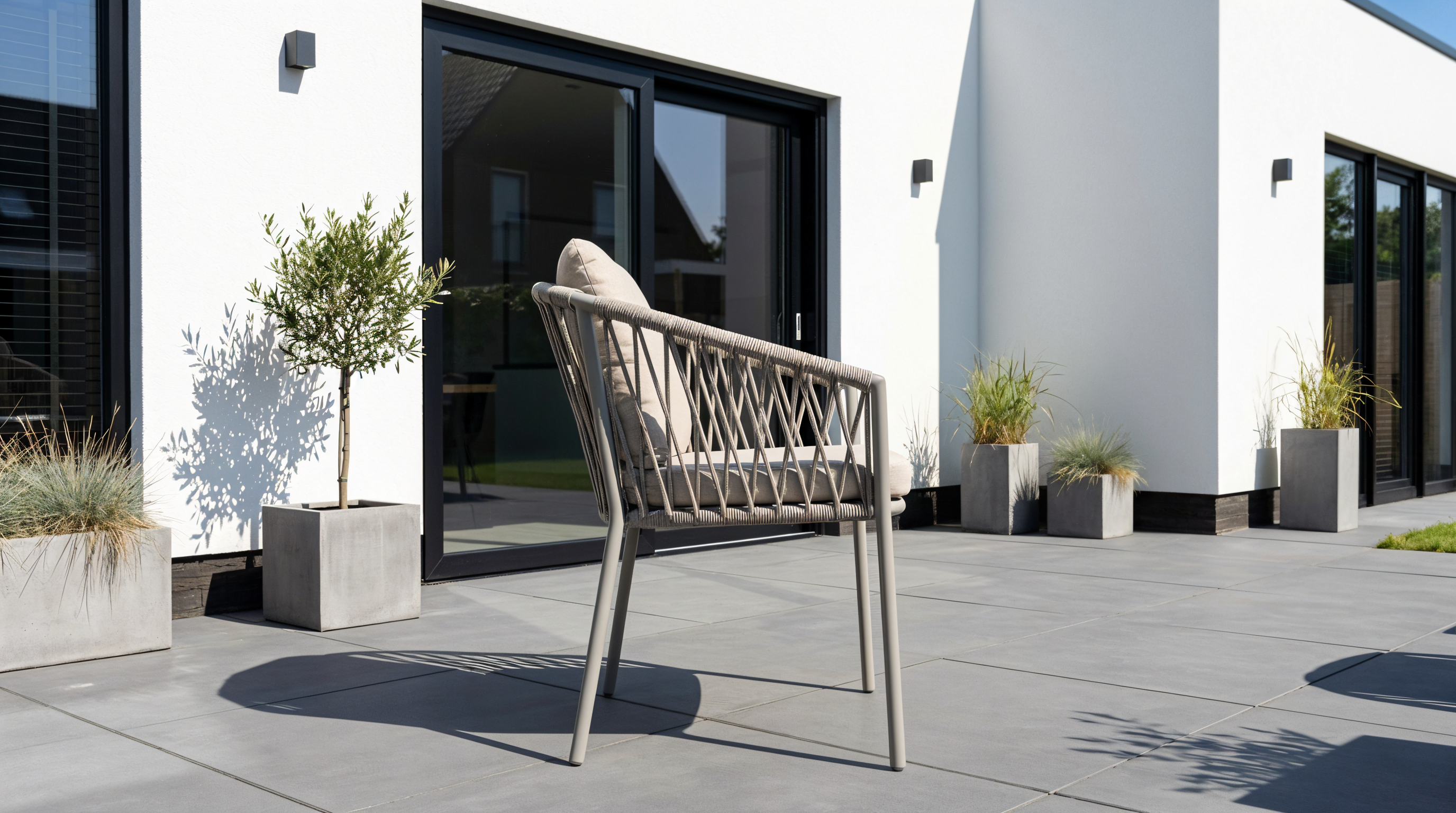Moderne dining tuinstoel met gevlochten rugleuning en licht kussen op een strak terras, voor een witte gevel met schuifpui en planten in potten.