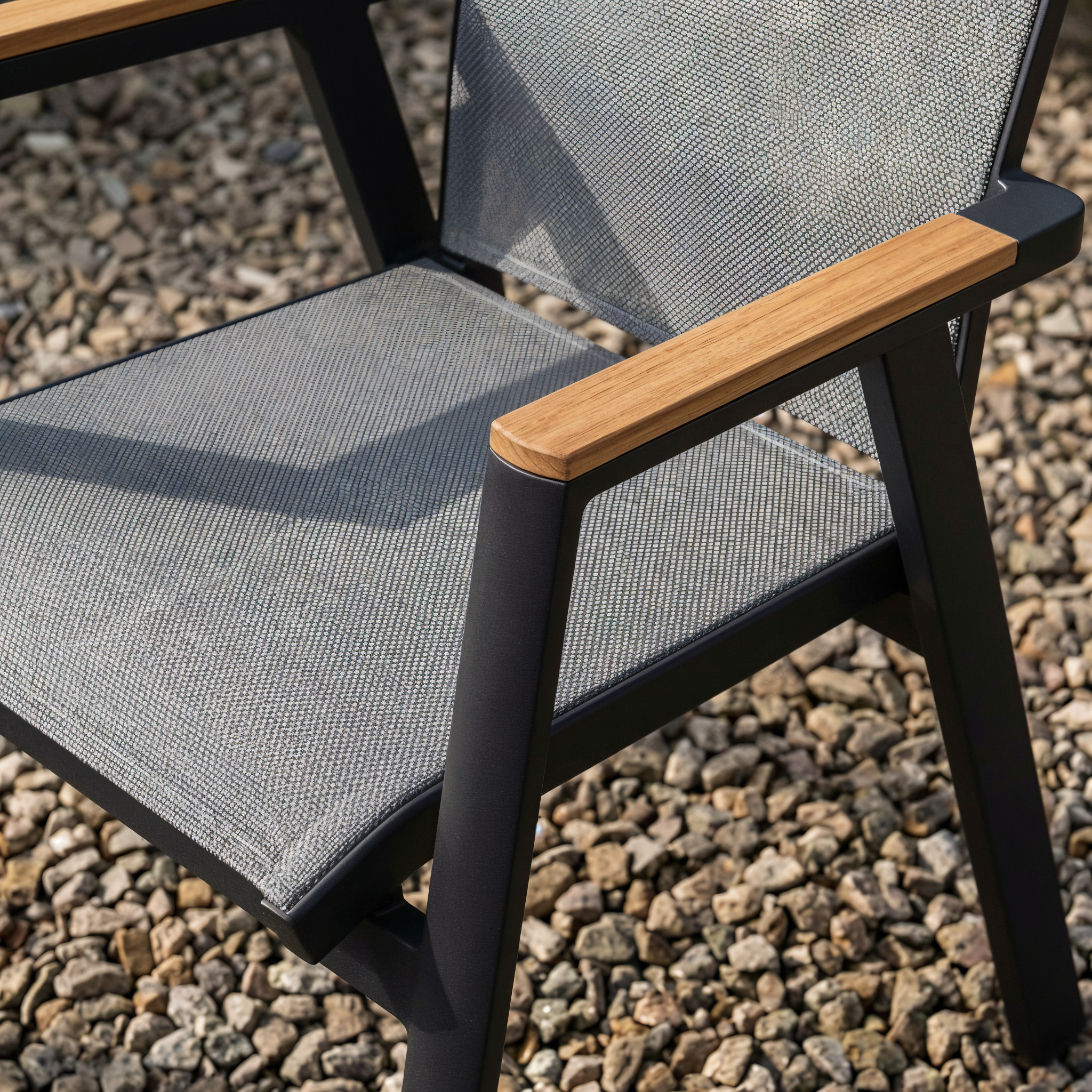 Detail van een tuinstoel met zwart aluminium frame, textilene zitting en houten armleuning op een grindterras.