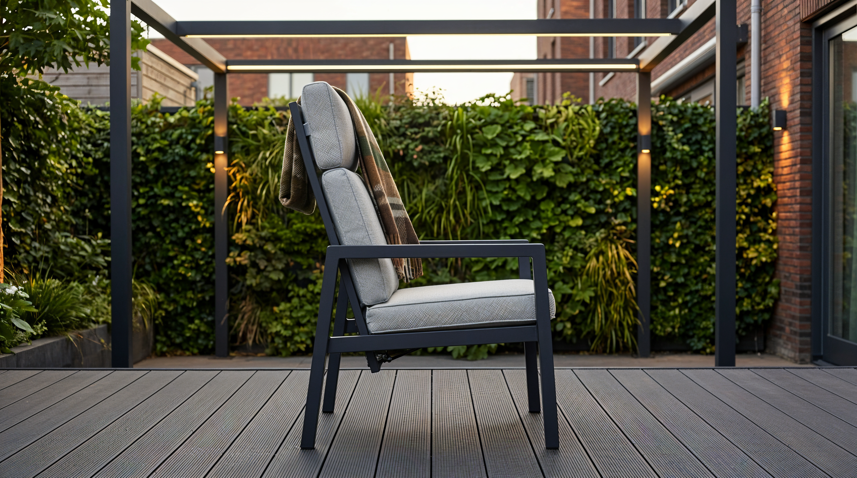 Verstelbare tuinstoel met hoge rugleuning, grijs zitkussen en zwart metalen frame op een terras met groene haag op de achtergrond.
