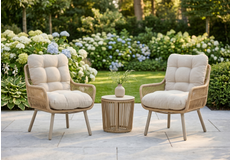 Set van twee luxe tuinstoelen met grijze kussens en blauwe sierkussens, naast een ronde wicker bijzettafel in een groene tuin