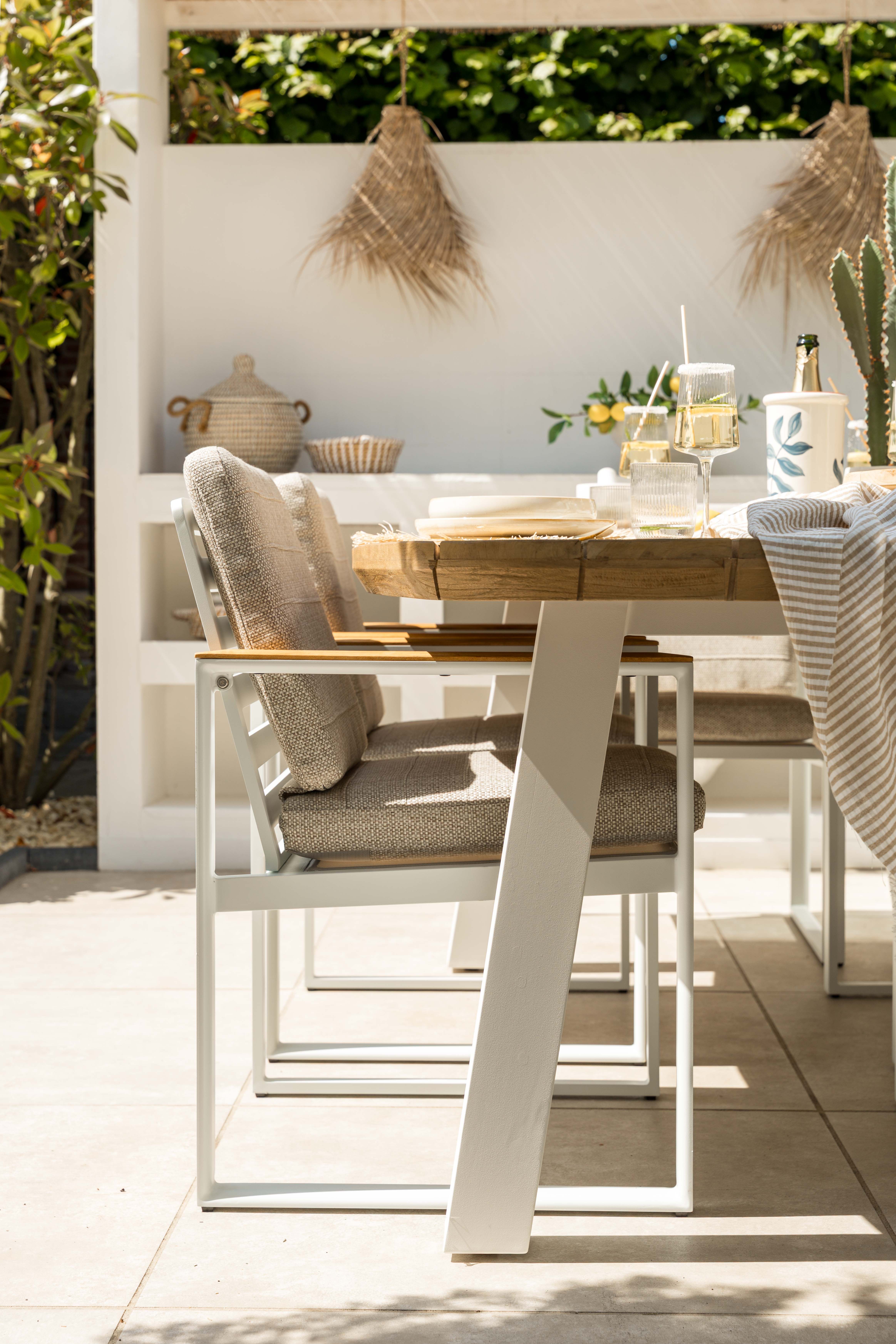 Ibiza stijl tuinset met houten eettafel en witte aluminium poten, comfortabele tuinstoelen met beige kussens en een sfeervol gedekte tafel op een modern terras.