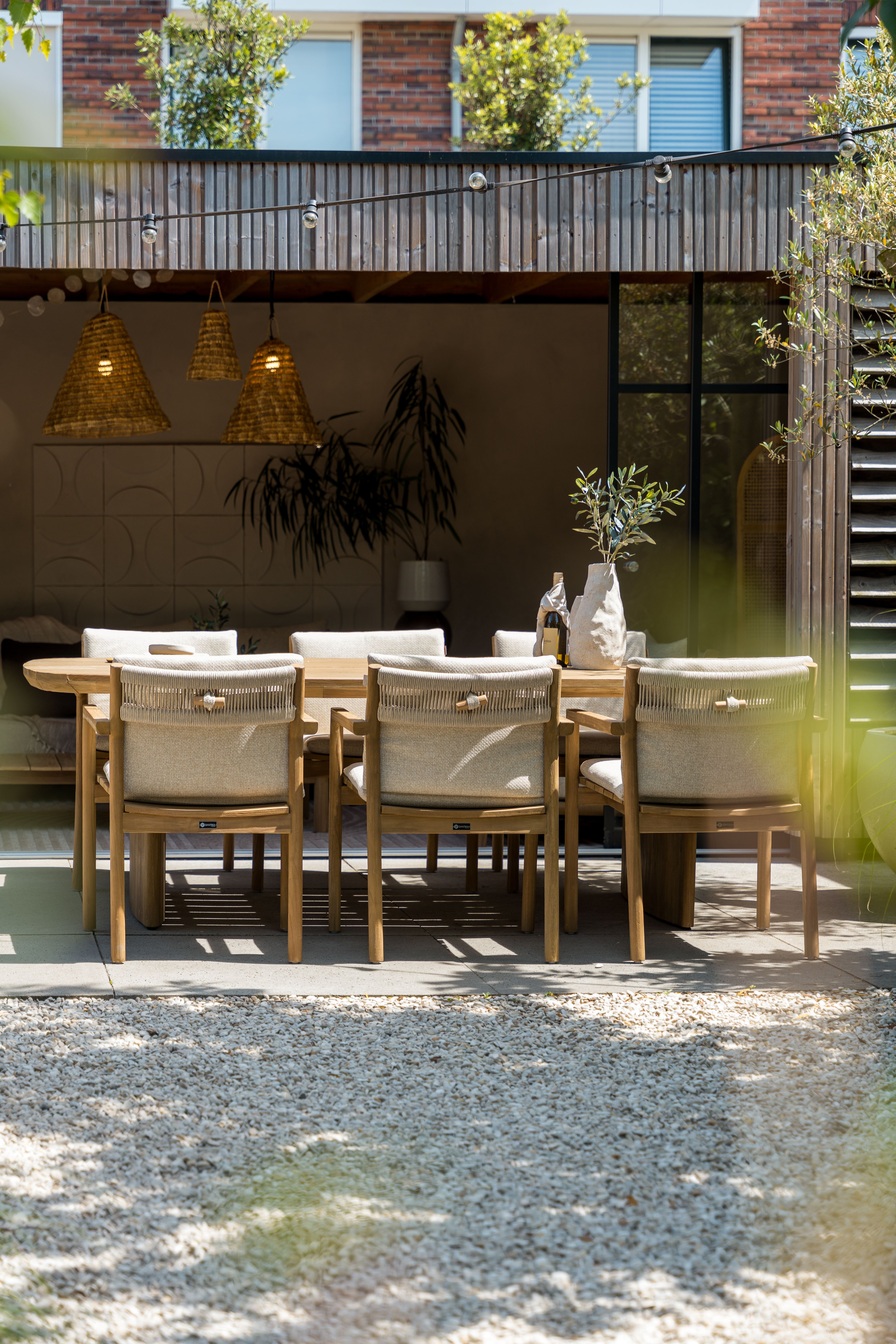 Japandi stijl tuinset met houten eettafel en acht bijpassende tuinstoelen met lichte kussens, sfeervol gedekt op een zonnig terras bij een moderne woning.