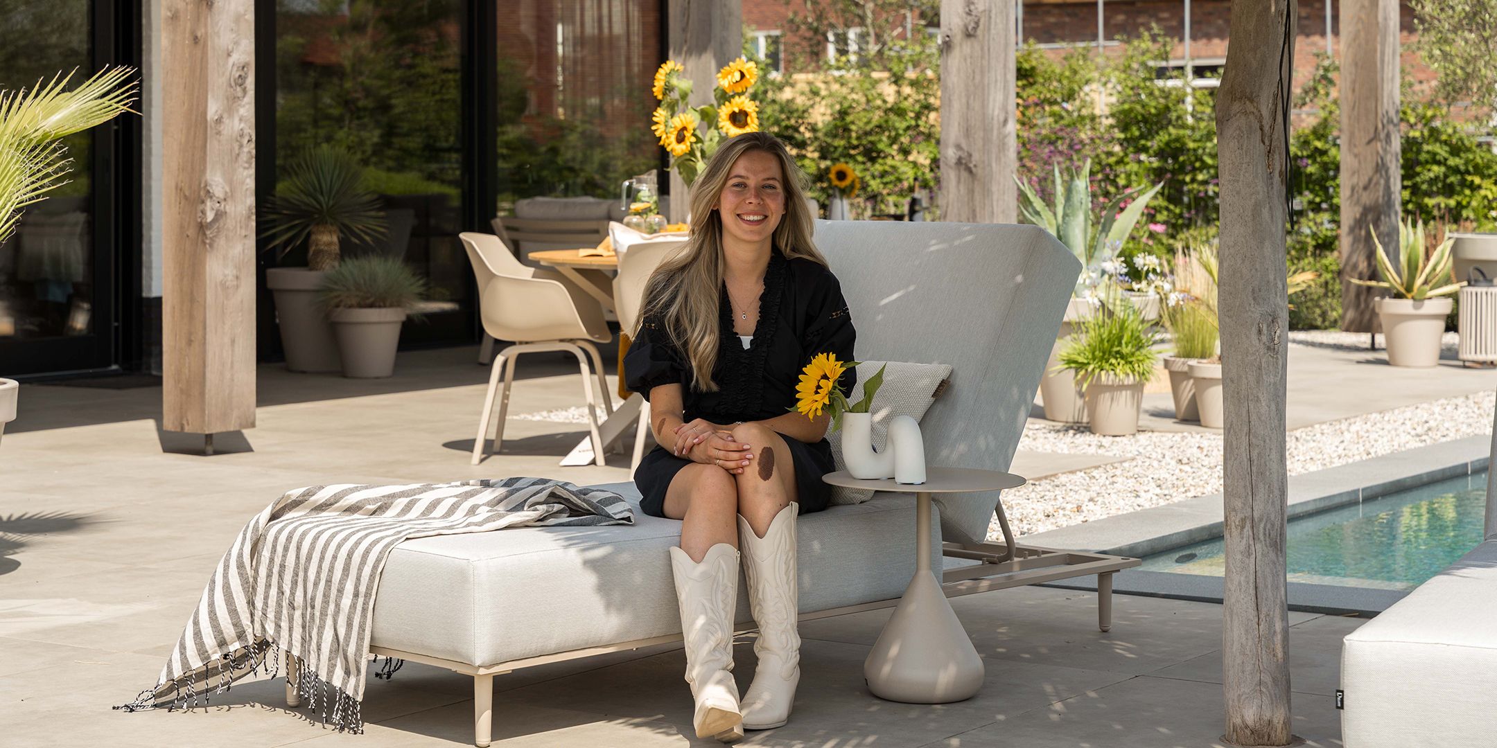 Vooraanzicht van onze winkel en tuinstyliste Hannah, met een geel geblokte plaid in haar hand naast een grijze loungebank.