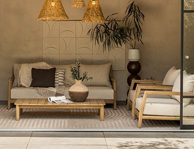 Vooraanzicht van een moderne loungeset met grijze tuinkussens en een houten loungetafel op een zonnig grindterras met groen.