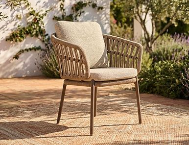 Vooraanzicht van een wicker tuinstoel met grijze tuinkussens op een grindterras naast een tuintafel in een landelijke tuin.