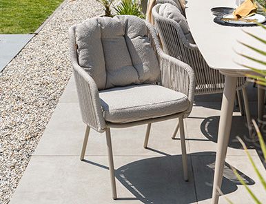 Vooraanzicht van een wicker tuinstoel met grijze tuinkussens op een grindterras naast een tuintafel in een landelijke tuin.