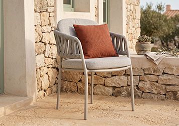 Vooraanzicht van een wicker tuinstoel met grijze tuinkussens op een grindterras naast een tuintafel in een landelijke tuin.